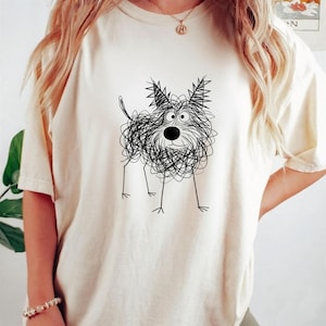 May include: Cream-colored t-shirt featuring a whimsical black and white scribble dog design. The dog has a round nose, big eyes, and spiky ears. The shirt is a relaxed fit, with short sleeves.
