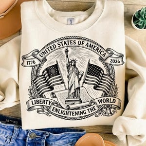 May include: Cream-colored sweatshirt featuring a black and white graphic of the Statue of Liberty, flags, and text. The text includes "United States of America," "Liberty," and "Enlightening the World." Dates 1776 and 2026 are also present.