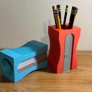 May include: Two pencil sharpeners, one red and one blue, with a pencil holder in the center. The pencil holder is red and has a silver metal blade. The sharpeners have a ribbed design and silver metal blades.