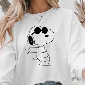 Puede incluir: Sudadera blanca de manga larga con una ilustraci&oacute;n en blanco y negro de Snoopy con gafas de sol. Snoopy hace una se&ntilde;al de pulgar hacia arriba y la palabra "cool" est&aacute; impresa en su camisa. Un estilo casual.
