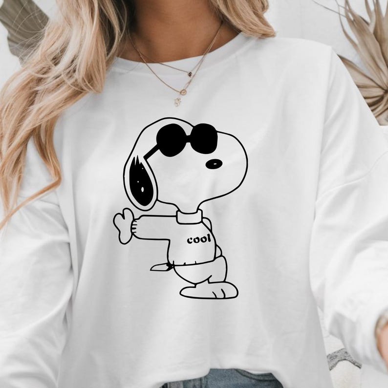 Puede incluir: Sudadera blanca de manga larga con una ilustraci&oacute;n en blanco y negro de Snoopy con gafas de sol. Snoopy hace una se&ntilde;al de pulgar hacia arriba y la palabra "cool" est&aacute; impresa en su camisa. Un estilo casual.