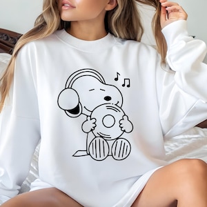 Puede incluir: Sudadera blanca con una ilustraci&oacute;n en blanco y negro de Snoopy con auriculares y sosteniendo un disco de vinilo. Notas musicales sobre la cabeza de Snoopy. Cuello redondo.