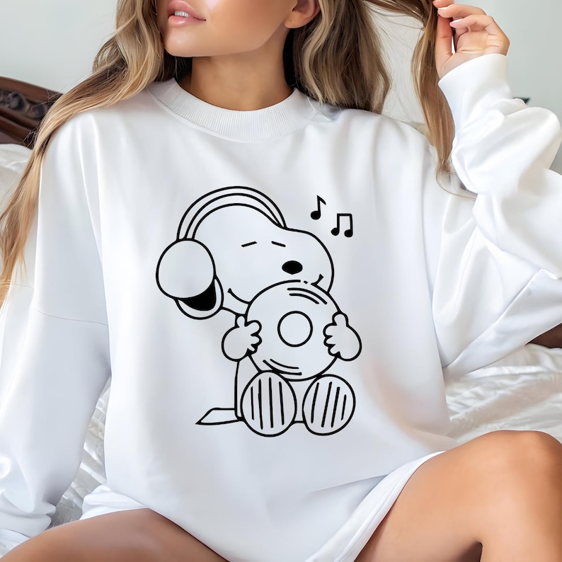 Puede incluir: Sudadera blanca con una ilustraci&oacute;n en blanco y negro de Snoopy con auriculares y sosteniendo un disco de vinilo. Notas musicales sobre la cabeza de Snoopy. Cuello redondo.