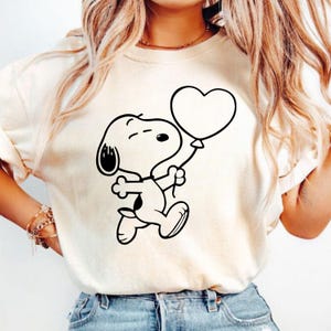 Puede incluir: Camiseta color crema con una ilustraci&oacute;n en blanco y negro de Snoopy sosteniendo un globo en forma de coraz&oacute;n. El personaje de dibujos animados se representa corriendo, con una expresi&oacute;n alegre. El dise&ntilde;o est&aacute; centrado en la parte delantera de la camiseta.