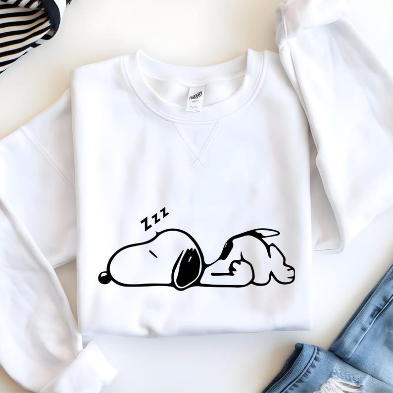 Puede incluir: Sudadera blanca de cuello redondo con un dibujo en blanco y negro de un perro de dibujos animados durmiendo y la palabra "Zzz". La sudadera es de material suave y tiene un corte cl&aacute;sico. El dise&ntilde;o es sencillo y juguet&oacute;n.