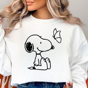 Puede incluir: Sudadera blanca con un dibujo en negro de Snoopy sentado y mirando una mariposa. El dise&ntilde;o es un simple dibujo lineal. La sudadera es de estilo casual y c&oacute;modo.