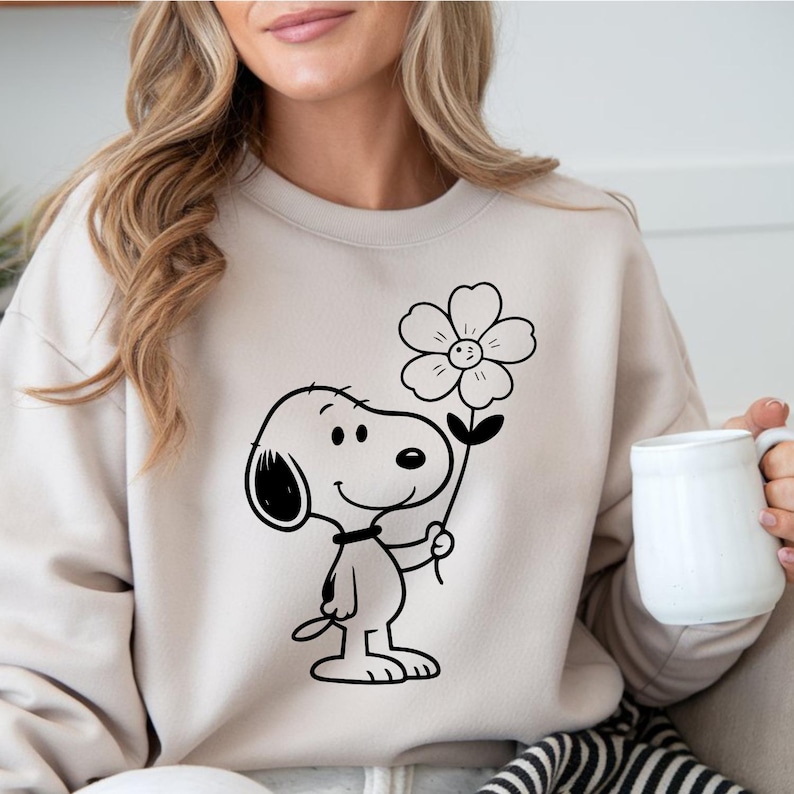 Puede incluir: Sudadera beige con un gr&aacute;fico en blanco y negro de Snoopy sosteniendo una flor. El personaje de dibujos animados est&aacute; sonriendo y la flor tiene cinco p&eacute;talos. La sudadera es de color neutro.