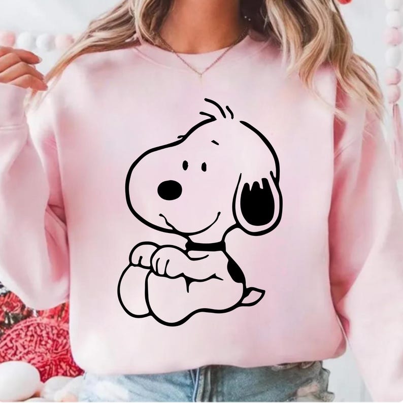 Puede incluir: Una sudadera rosa claro con una ilustraci&oacute;n de dibujos animados en blanco y negro de un perro. El perro est&aacute; sentado con las patas juntas y tiene una nariz, orejas y collar negros. La sudadera es de color liso.