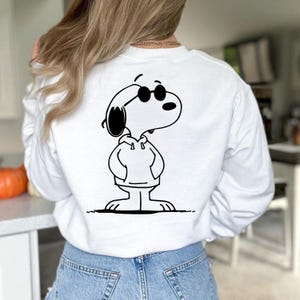 Puede incluir: Sudadera blanca con una ilustraci&oacute;n en blanco y negro de Snoopy con gafas de sol y una sudadera con capucha. El personaje de dibujos animados est&aacute; de pie con las manos en los bolsillos. La sudadera es una parte superior informal de manga larga.