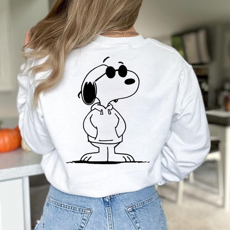 Puede incluir: Sudadera blanca con una ilustraci&oacute;n en blanco y negro de Snoopy con gafas de sol y una sudadera con capucha. El personaje de dibujos animados est&aacute; de pie con las manos en los bolsillos. La sudadera es una parte superior informal de manga larga.