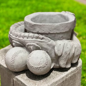 May include: A gray concrete baseball-themed planter. The planter is shaped like a baseball glove holding two baseballs. The top of the glove forms a circular opening for planting. The planter is sitting on a gray stone base.