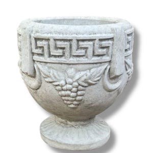 May include: A gray, weathered-looking urn with a pedestal base. The urn features a Greek key design around the top and a grape cluster motif. The vessel is likely made of stone or concrete.
