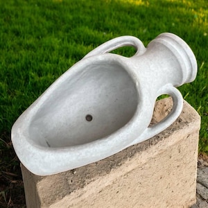 May include: A gray, weathered-look planter in the shape of an amphora. The planter has two handles and a small drainage hole. It is sitting on a concrete block, with green grass in the background.