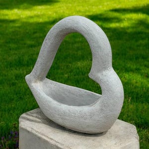 May include: A light gray, abstract sculpture with a curved, handle-like form. The sculpture sits on a concrete base against a backdrop of green grass. The artwork has a textured surface and a modern aesthetic.
