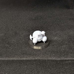 May include: A small, white, 3D-printed figurine of a cartoonish character with a round body, two large eyes, and small appendages. The figure is mounted on a dark gray circular base, set against a dark background.