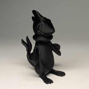 May include: A black 3D-printed figurine of a cartoonish creature with a seal-like body, flipper-like arms, and a pointed head. The figure has a collar around its neck and is posed with its arms out. The figurine is approximately 4 inches tall.