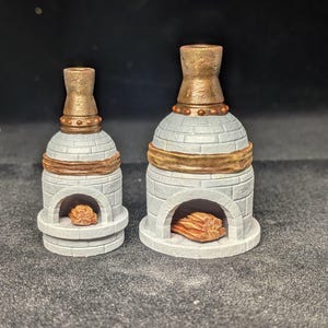 May include: Two miniature, decorative ovens. The ovens are white with a brick pattern, a gold band, and a gold top. The oven openings contain a brown, wood-like material. One oven is smaller than the other.