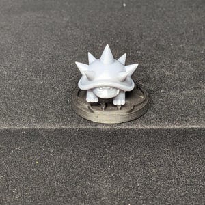 May include: A light gray, spiky turtle figurine. The turtle has a textured shell with multiple sharp spikes. It is standing on a dark gray, circular base. The figurine is a 3D-printed item.