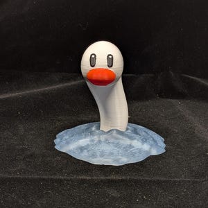 May include: A 3D printed figurine of a white, worm-like creature with a rounded head, black oval eyes, and a bright orange beak. The creature emerges from a translucent, light blue textured base that mimics the appearance of rippling water.