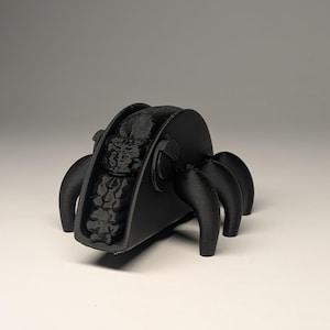 May include: A black 3D-printed taco holder with six curved legs. The taco holder is designed to resemble a spider, with the taco shell forming the body and the legs providing support. The interior of the taco holder is textured.