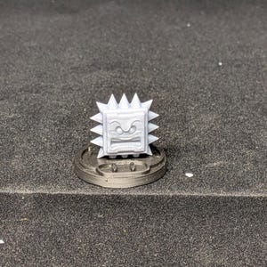 May include: A gray 3D-printed Thwomp figure, a character from the Super Mario video game series. The figure has a square body with a frowning face and spiky edges, mounted on a circular base.