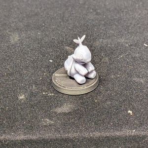 May include: A small, light purple figurine of a cartoon character with a large head, sitting on a circular base. The figure has a backpack and a leaf-like design on its head. The base is dark gray with a textured surface.