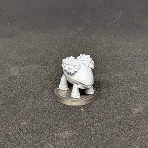 May include: A small, gray figurine of a creature with a rounded body, four legs, and two fluffy, textured elements on its back. The figurine is mounted on a small, circular base. The background is a dark, textured surface.