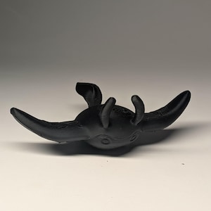 May include: A black, stylized sculpture of a manta ray. The sculpture has a textured surface and is set against a white background. The ray's wings are spread wide, and it has two small appendages near its head.