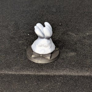 May include: A small, light gray figurine of a character with a rounded body, two large eyes, and a head shaped like a hand with five fingers. The figurine sits on a dark gray, circular base with a geometric design.