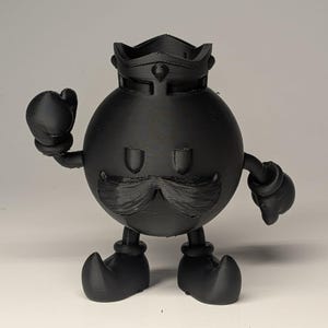 May include: A black 3D-printed figurine of a cartoon character with a crown, mustache, and boxing gloves. The figure has a round body, small feet, and is posed with one arm raised. The figurine is made of a matte black material.