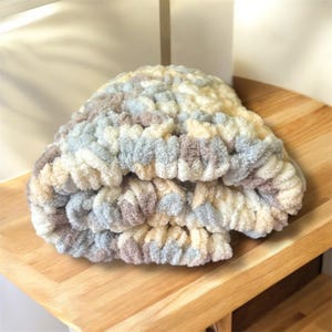 May include: A soft, chunky knit hat in a neutral color palette of cream, light blue, and taupe. The hat is made of a thick, textured yarn, and appears to be folded over. The hat is sitting on a wooden surface.