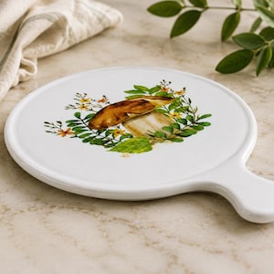May include: A round white ceramic serving board with a handle, featuring a central illustration of a brown mushroom surrounded by green leaves and small yellow flowers. The surface is smooth and glossy, showcasing a clean, minimalist design.