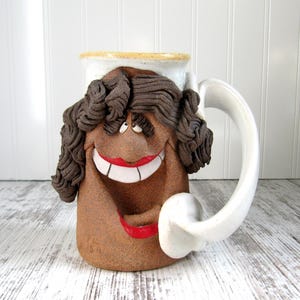 May include: A whimsical ceramic mug with a smiling face design. The mug features a brown face with dark brown curly hair, red lips, and white teeth. The handle is white, and the rim is a light beige color.