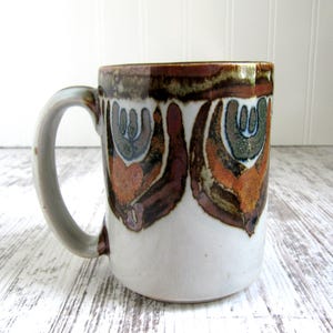 May include: A ceramic mug with a white base and a brown, orange, and blue abstract design. The mug has a light gray handle and a brown rim. The design features stylized shapes and patterns.