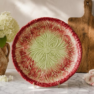 May include: A round ceramic plate featuring a central embossed light green leaf pattern surrounded by a textured, deep red rim. The plate has a scalloped, uneven edge and a glossy finish, showcasing a vibrant contrast between the green center and red border.