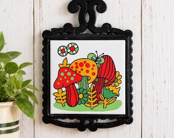 May include: A square ceramic tile featuring a colorful graphic illustration of red, yellow, and orange mushrooms with green stems, surrounded by stylized flowers and a butterfly, set within a decorative black metal frame with a hanging loop.