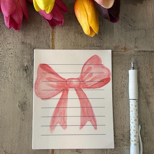May include: A white notepad with lined paper and a pink watercolor bow illustration. The notepad is on a wooden surface with a pen next to it.