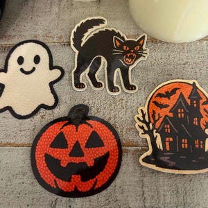May include: A collection of Halloween-themed stickers. Includes a smiling ghost, a black cat with orange eyes, a jack-o'-lantern, and a haunted house with bats. The stickers have a textured appearance and are set against a light gray background.