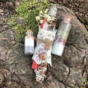 Könnte beinhalten: Eine Auswahl an spirituellen Gegenständen, darunter ein Salbeibündel, das in Spitze und Bindfaden gewickelt ist, zwei kleine Glasflaschen mit Korken und eine rote Kerze. Die Gegenstände sind auf einer strukturierten, braunen Oberfläche mit grünem Moos angeordnet.