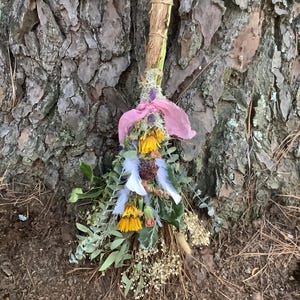 Puede incluir: Un manojo de salvia hecho a mano con flores amarillas secas, plumas blancas y follaje verde, atado con una cinta rosa. El paquete está hecho con materiales naturales y está contra un fondo de corteza de árbol texturizada.