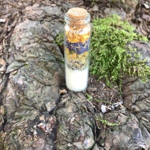May include: A small glass bottle filled with layers of colorful dried flowers and herbs, topped with a cork stopper. The layers include white, yellow, blue, and purple elements. The bottle is set on a textured, brown, and green natural surface.