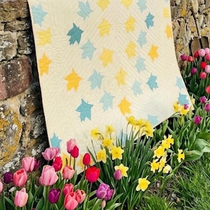 May include: A cream-colored quilt with a leaf pattern in yellow and teal blue rests on a stone wall. The quilt is surrounded by a vibrant flower bed of tulips and daffodils.