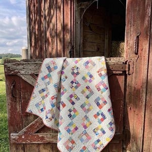 May include: A patchwork quilt with a white base and vibrant, multi-coloured squares. The quilt is displayed over a weathered, red barn door, creating a rustic, vintage feel. The quilt has a pink border around its edges.