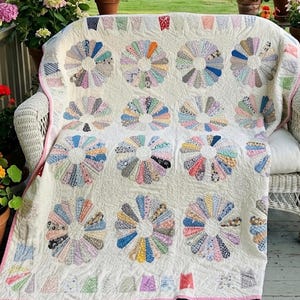 May include: A cream-colored quilt featuring a repeating pattern of Dresden plate flower motifs. Each flower is composed of multi-colored, patterned fabric petals arranged in a circular design. The quilt has a pink border and intricate stitching.