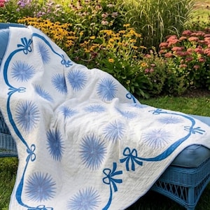 May include: A white quilt featuring a repeating pattern of blue starburst or flower-like motifs. The quilt is bordered by a blue ribbon design that includes several tied bows at intervals along the edges of the fabric.