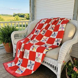 May include: A vibrant red and white patchwork quilt with star patterns, draped over a white wicker loveseat. The quilt features red squares and white squares with colourful star-shaped designs. The scene is set on a wooden porch with potted plants.