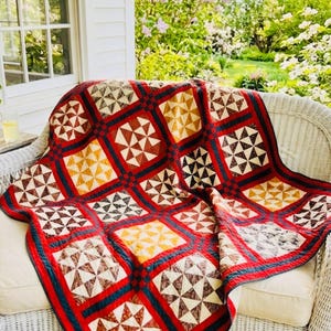May include: A colorful quilt with a geometric pattern of triangles and squares in shades of red, brown, yellow, and white. The quilt is draped over a white wicker chair on a porch, with a view of a garden.