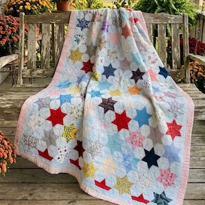 May include: A quilt featuring a repeating star pattern made from various multi-colored fabric scraps. The stars are set against a light blue background, and the entire piece is finished with a solid light pink border along the edges.