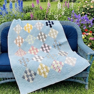 May include: A light blue quilt featuring a patchwork design of diamond-shaped blocks. Each block contains a unique pattern in colors including yellow, pink, white, and dark blue. The quilt is stitched with a linear pattern throughout the fabric.