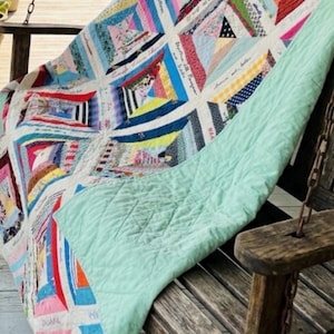 May include: A colorful patchwork quilt with a light green backing rests on a wooden porch swing. The quilt features a variety of fabric patterns and colors, including pink, blue, and red. Some quilt blocks have handwritten text.